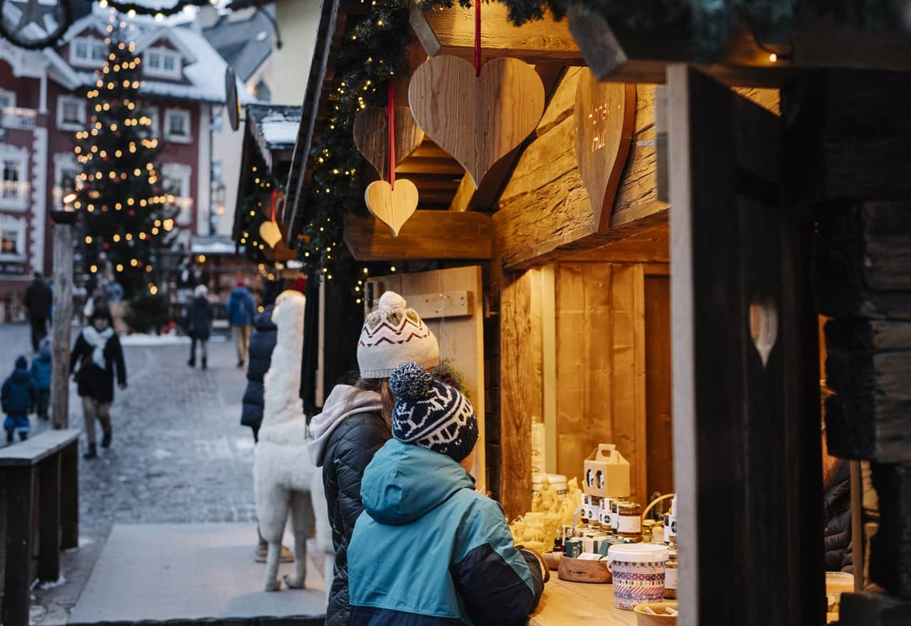 Persone con cappelli invernali osservano prodotti a una bancarella di legno decorata con cuori e luci natalizie