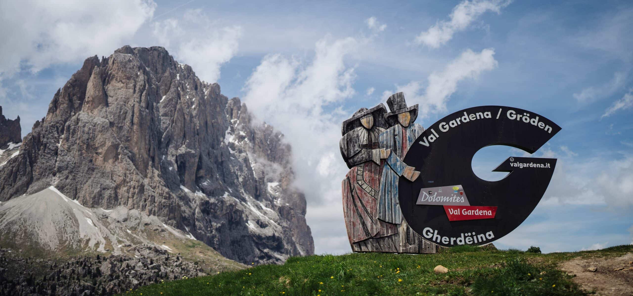 Scultura lignea e cartello Val Gardena / Gröden su prato, parete rocciosa dolomitica sullo sfondo