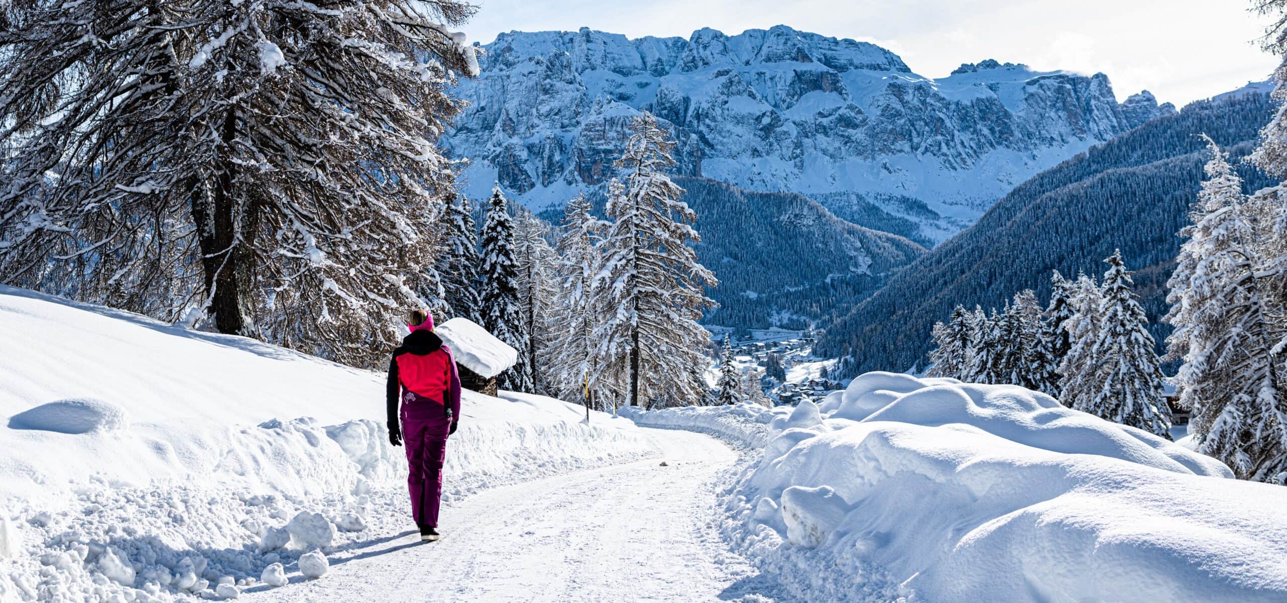 Persona con giacca rosa cammina su strada innevata verso una valle tra alberi e montagne innevate