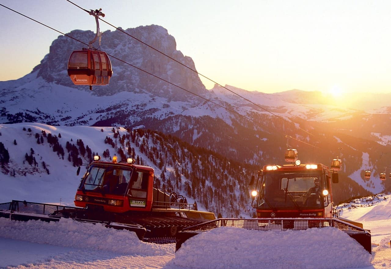 Due battipista sul pendio innevato, cabine della funivia in quota e montagne al tramonto