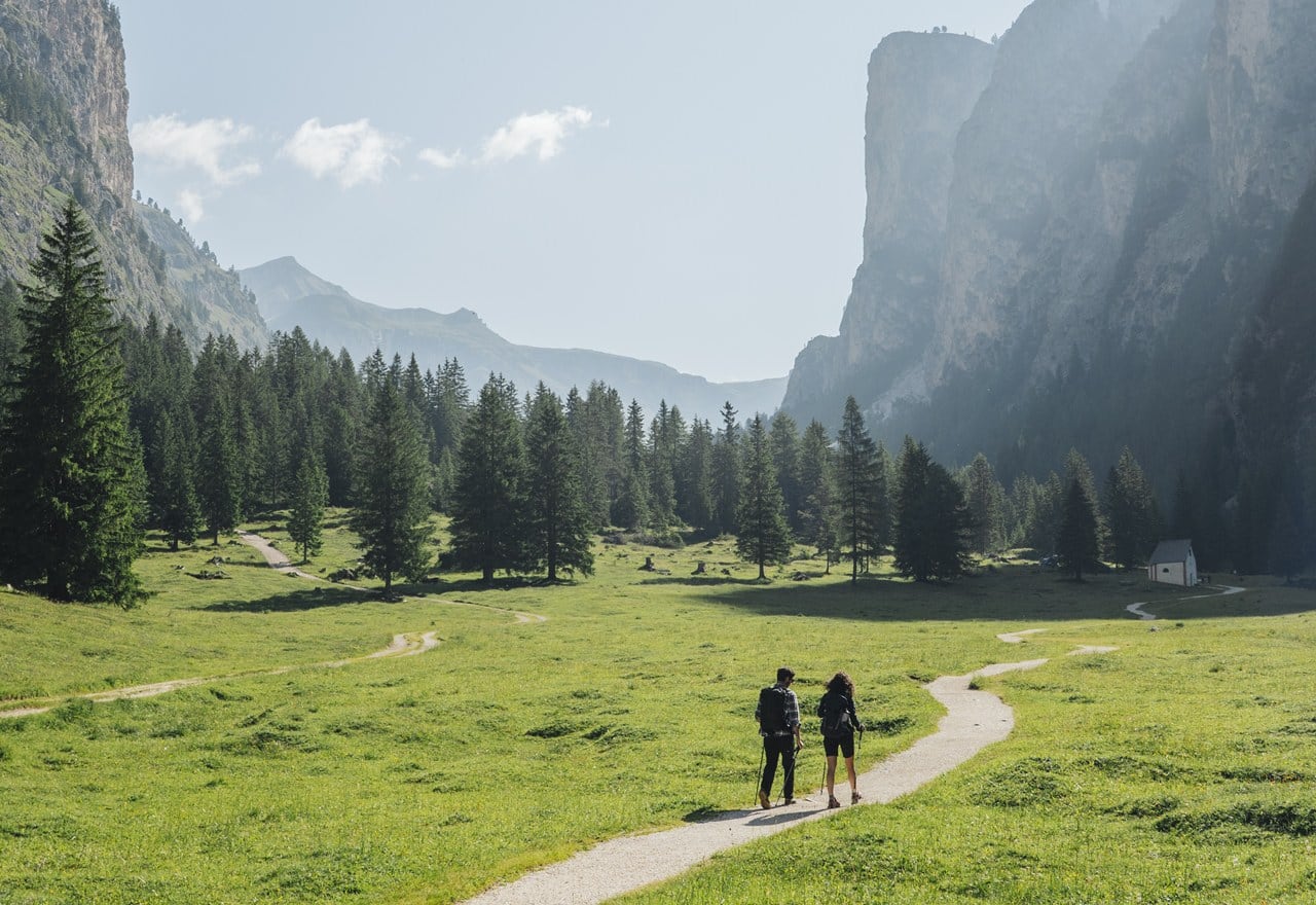 Due escursionisti con zaini percorrono un sentiero nel prato verso un bosco e alte pareti rocciose.