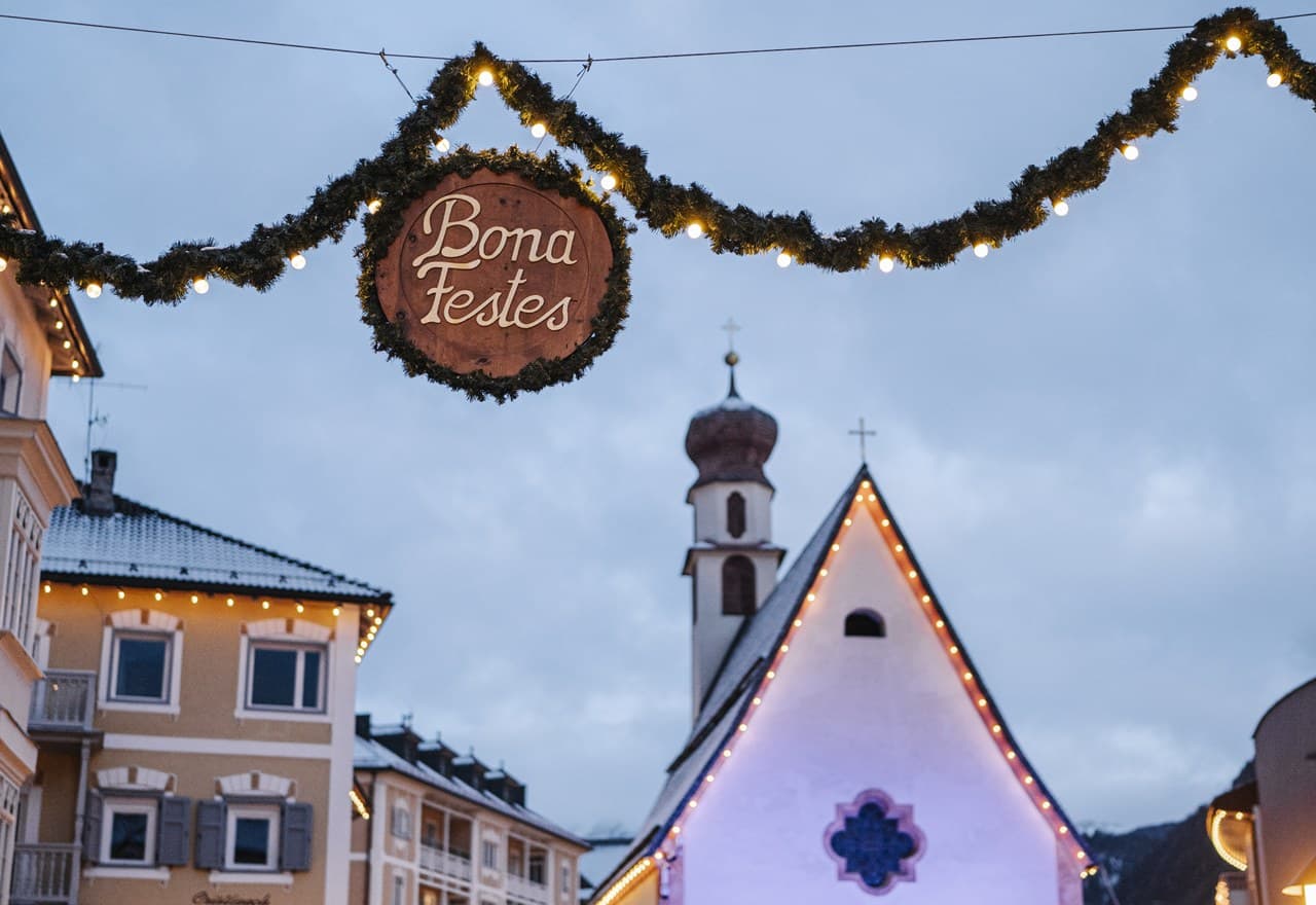 Targa circolare Bona Festes sospesa a festone luminoso su strada con chiesa e case innevate