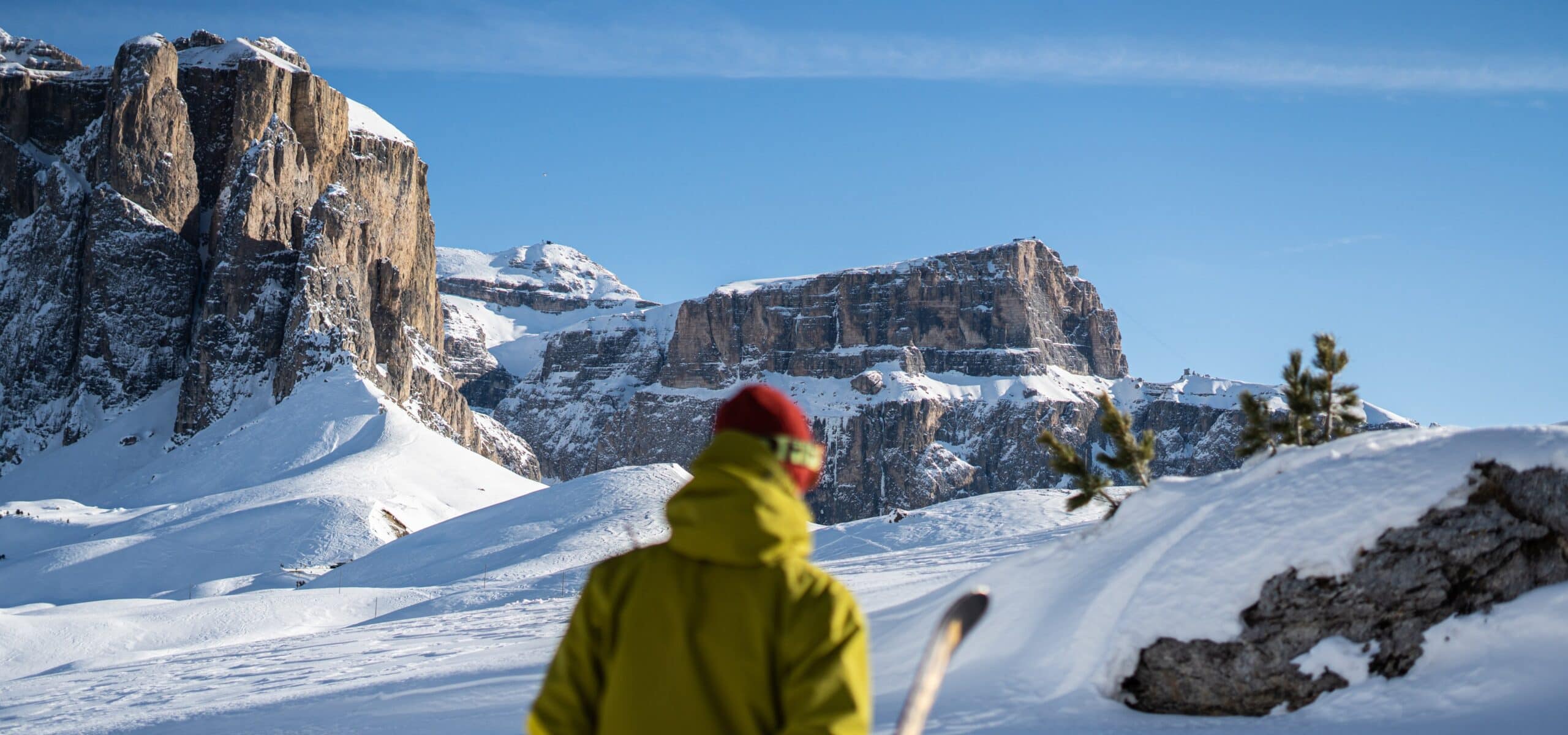 Persona con giacca gialla, berretto rosso e gli sci, vista da dietro verso pareti rocciose innevate