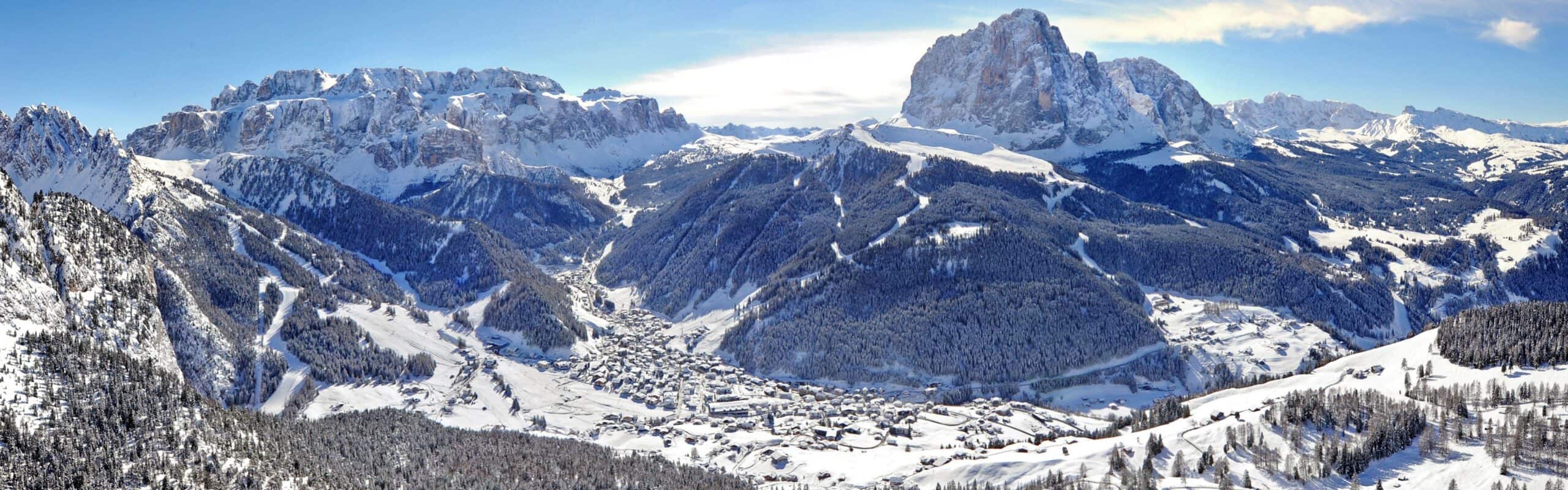 Valle innevata con paese in centro, piste da sci e foreste, massiccio roccioso sullo sfondo