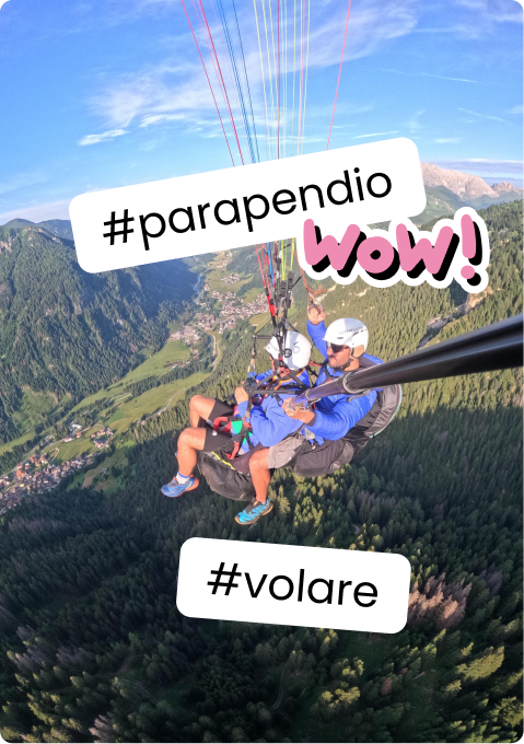 Due persone in tandem su parapendio, elmetti e selfie con bastone sopra valle montana; sticker '#parapendio' '#volare'
