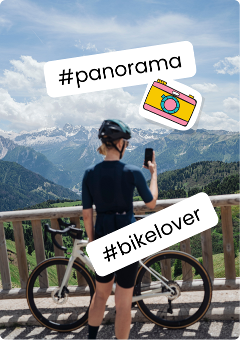 Ciclista di spalle con casco e bici che scatta col cellulare da un balcone con vista sulle montagne.