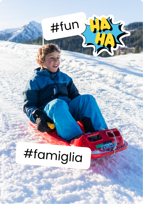 Bambino sorridente su slitta rossa che scivola sulla neve, montagne innevate sullo sfondo