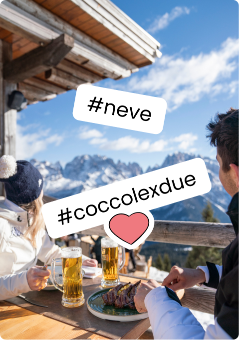 Due persone sedute a un tavolo su terrazza di montagna, birre e piatto di carne, montagne innevate sullo sfondo