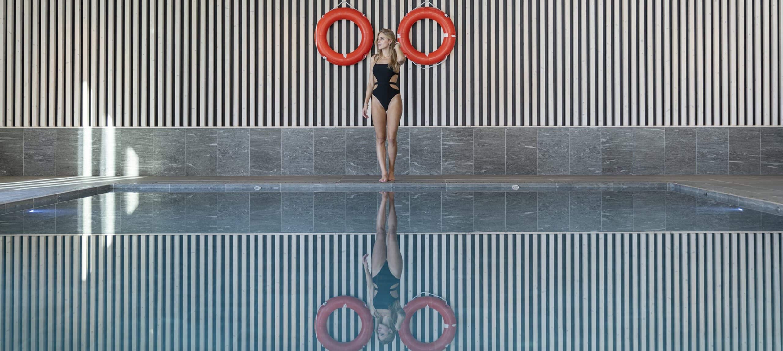 Donna in costume nero in piedi al bordo di una piscina interna con due salvagenti rossi sul muro e riflesso