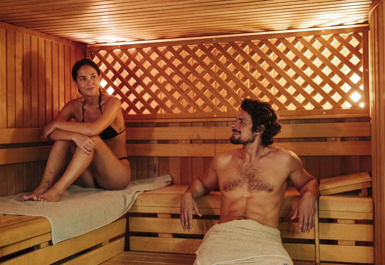 Donna in bikini seduta su panca e uomo con asciugamano alla vita in sauna di legno