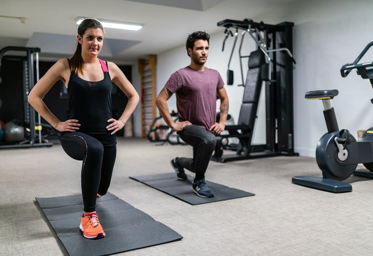 Due persone eseguono affondi su tappetini in una palestra con attrezzi e cyclette sullo sfondo