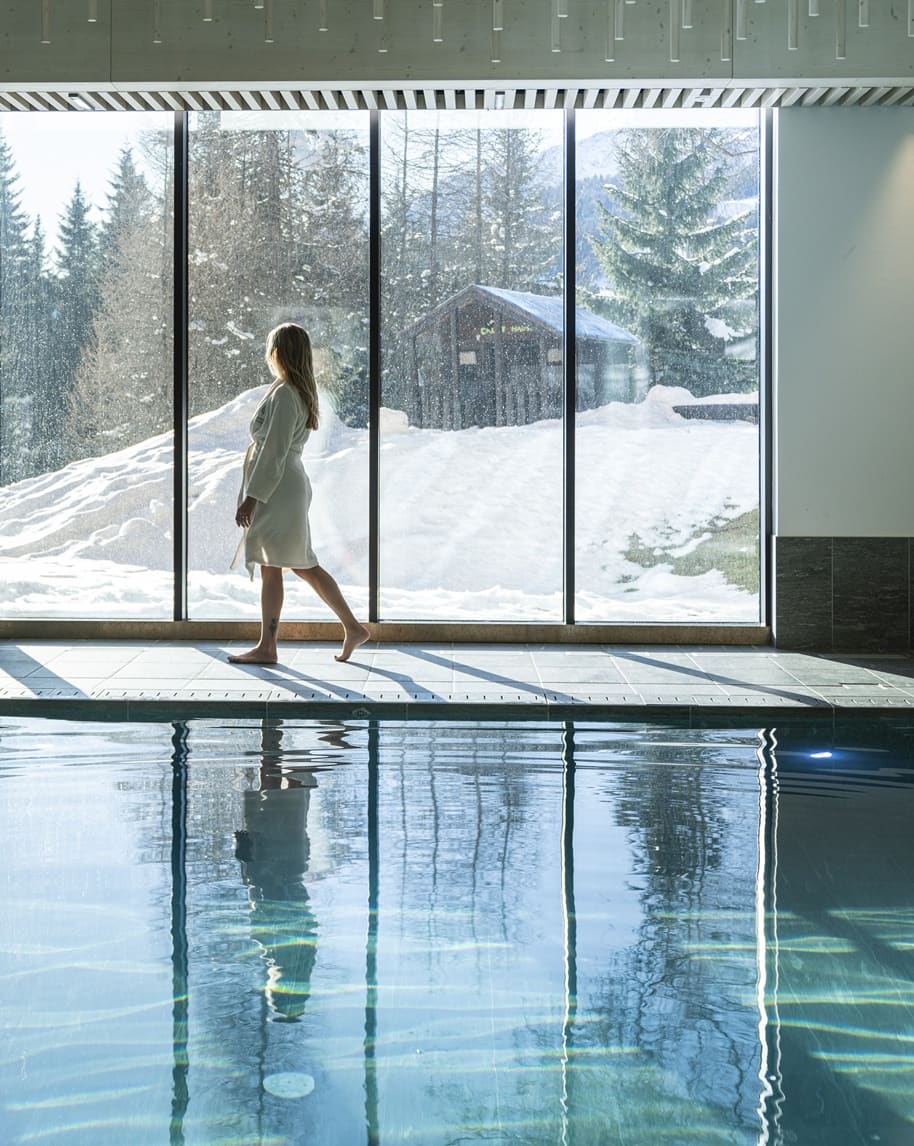 Donna in accappatoio cammina a piedi nudi lungo la piscina interna con grandi vetrate su paesaggio innevato
