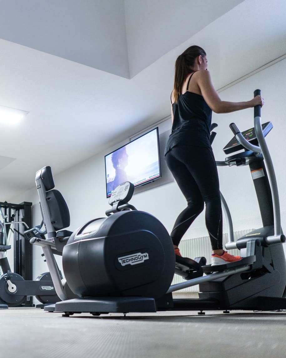 Donna su ellittica in palestra, vista laterale, altre attrezzature e schermo TV sullo sfondo