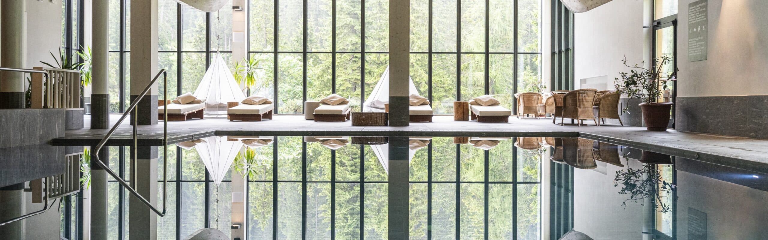 Piscina interna con vetrate panoramiche, lettini e poltrone lungo il bordo; foresta riflessa sull'acqua