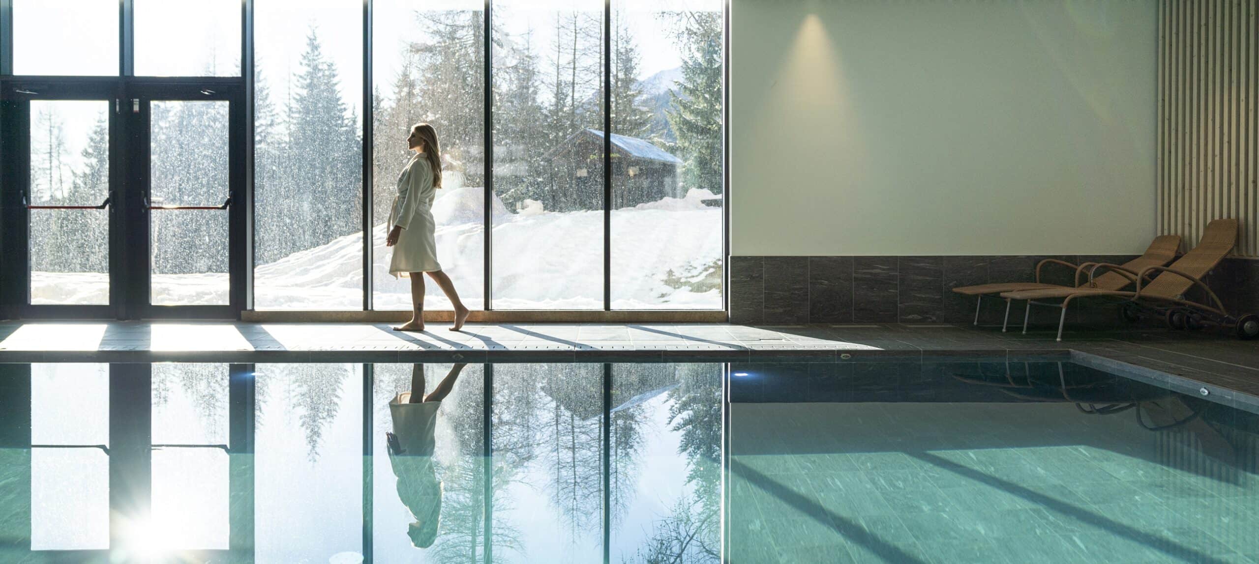 Donna in accappatoio cammina a bordo piscina davanti a vetrata con paesaggio innevato, riflesso nell'acqua