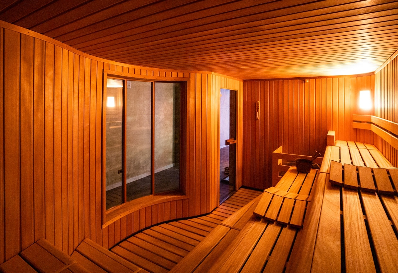 Sauna in legno con panche a più livelli, finestra curva e porta verso corridoio, luce calda