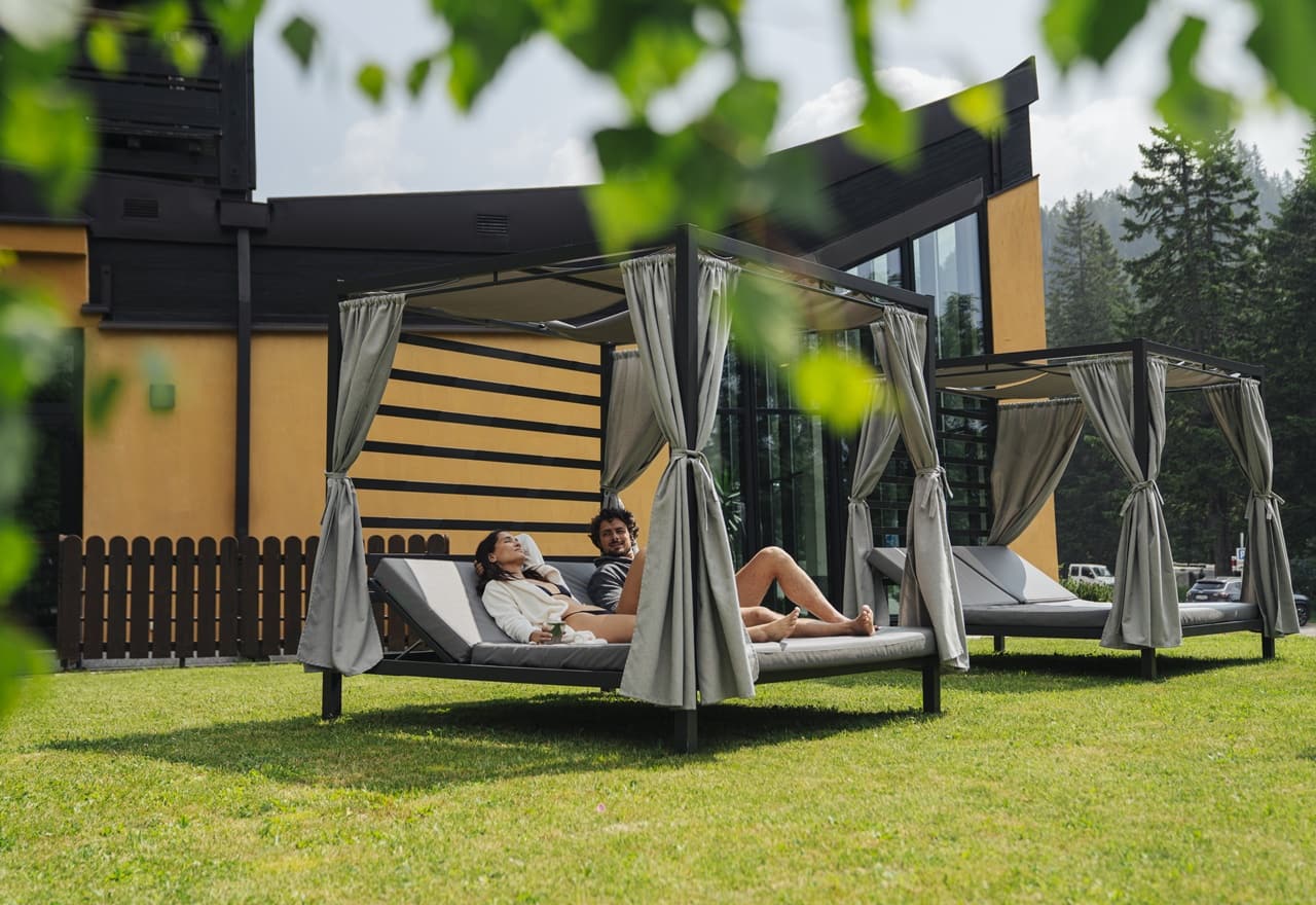 Coppia sdraiata su lettini coperti in giardino davanti a un edificio moderno con pini sullo sfondo
