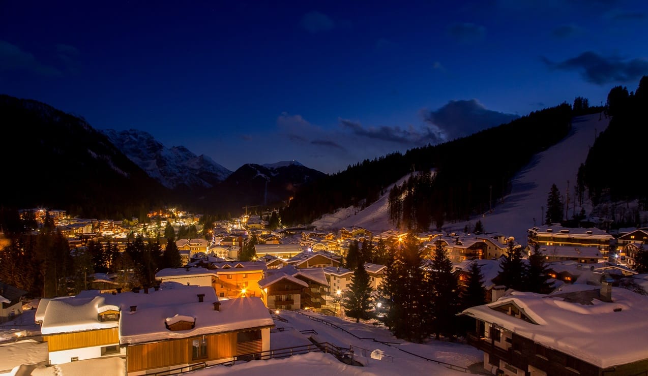 Paese montano innevato di sera, case illuminate, pista da sci sul pendio e montagne sullo sfondo