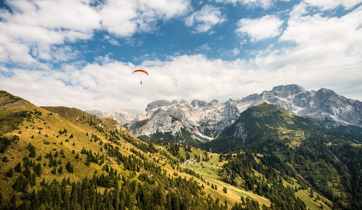 Parapendio arancione sorvola vallata erbosa con pendii boscosi e massiccio roccioso sullo sfondo.