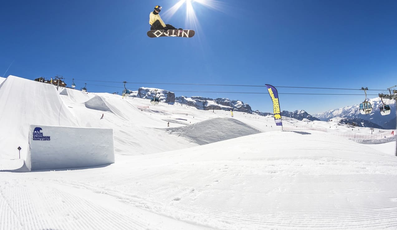 Snowboarder in aria sopra ostacoli del Ursus Snowpark, piste innevate, cabine panoramiche e montagne sullo sfondo
