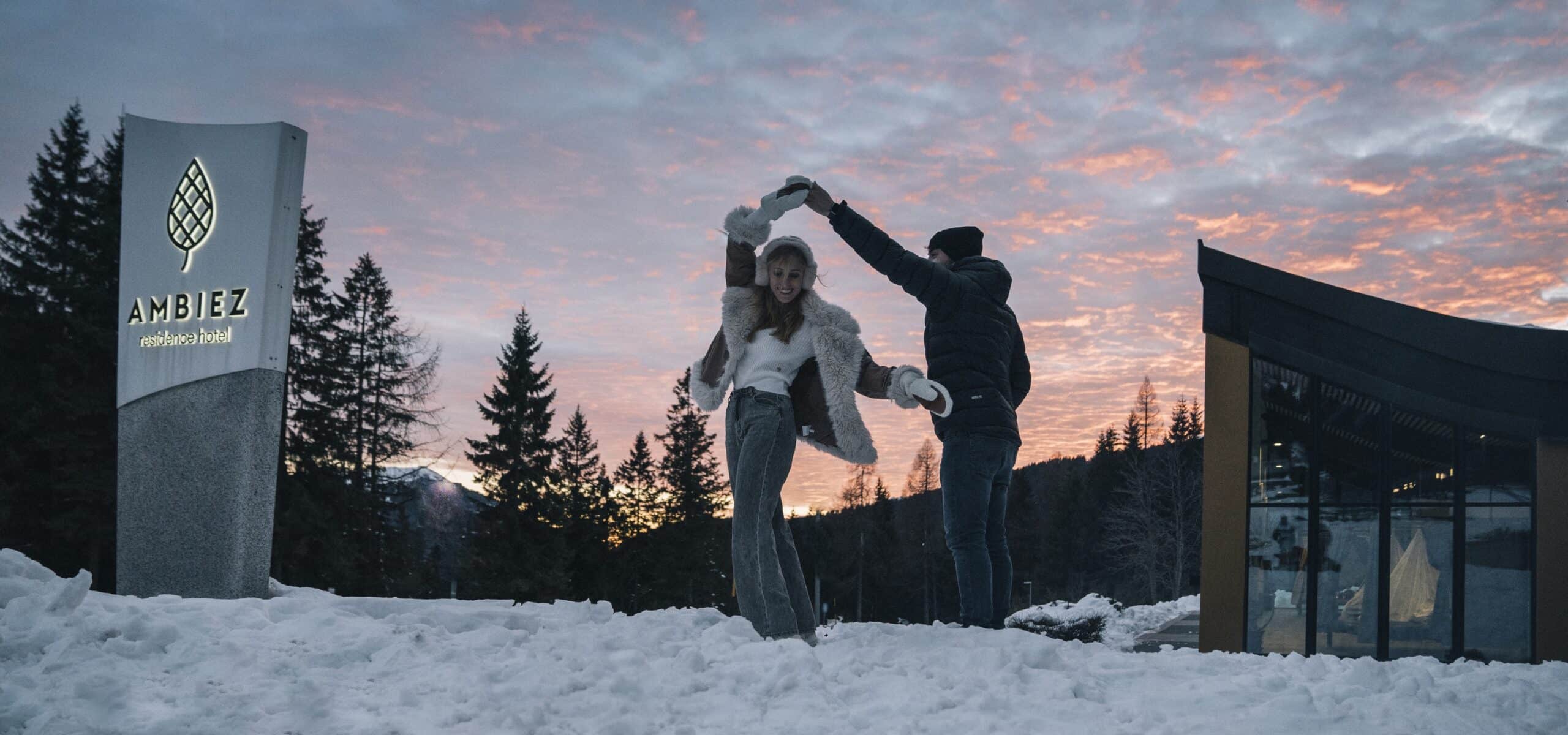 Coppia che balla sulla neve al tramonto davanti allinsegna AMBIEZ residence hotel e a un edificio vetrato