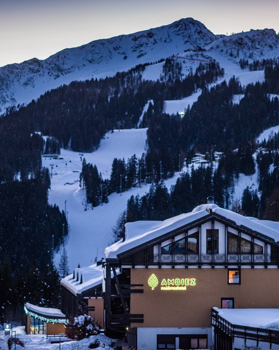 Edificio di Ambiez Residencehotel innevato in primo piano, piste da sci e montagna sullo sfondo