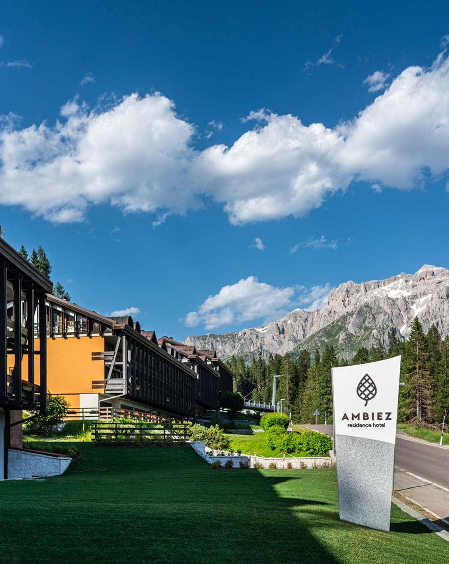 Edificio di Ambiez Residencehotel su prato, con alberi e montagne rocciose sotto cielo azzurro e nuvole.
