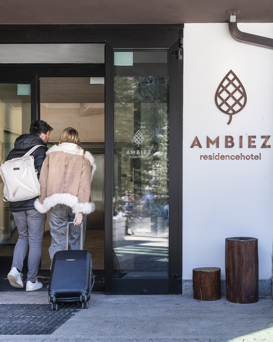 Coppia con valigia entra nell'edificio di Ambiez residencehotel, ingresso con porta a vetri