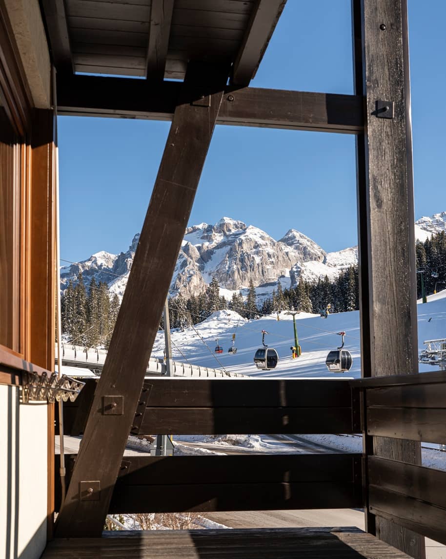 Balcone in legno che incornicia montagne innevate e cabine della funivia sopra una pista da sci