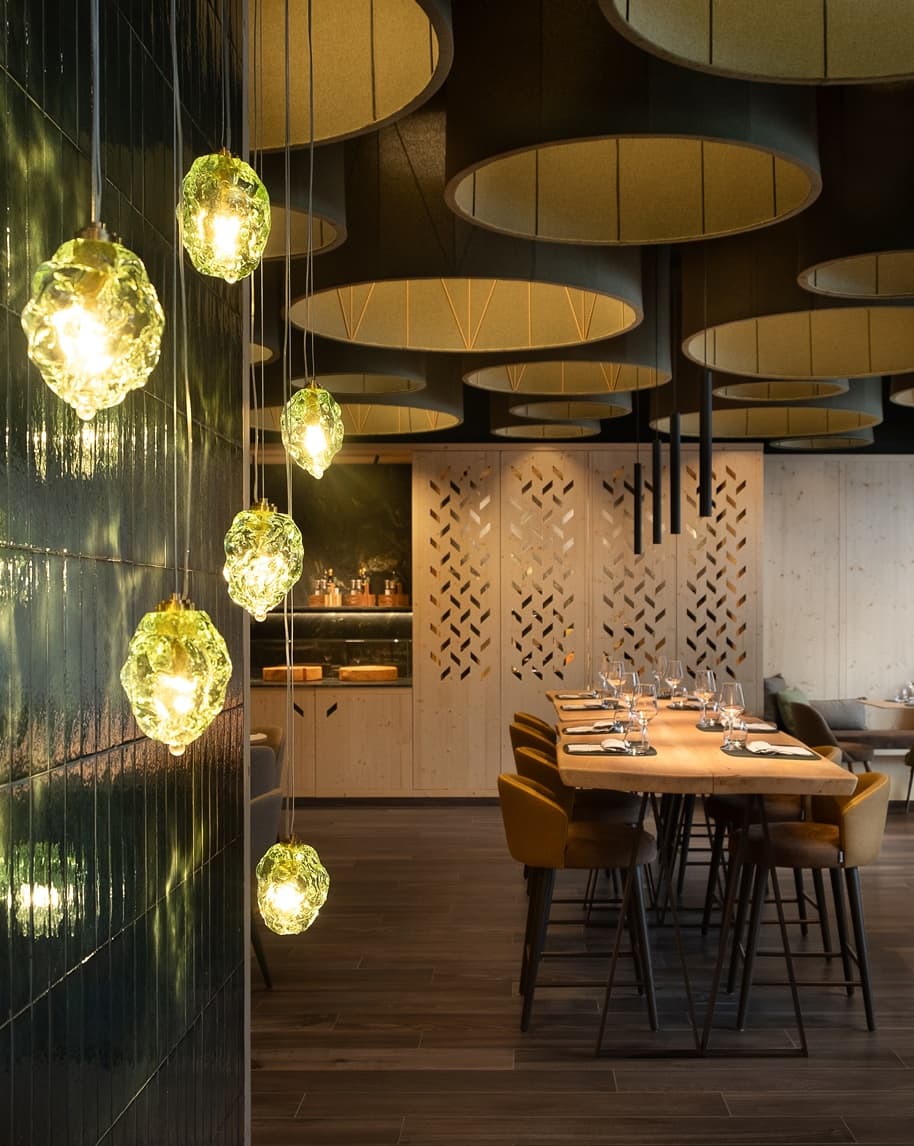 Lampade pendenti in vetro verde; tavolo lungo apparecchiato con sedie in ristorante moderno
