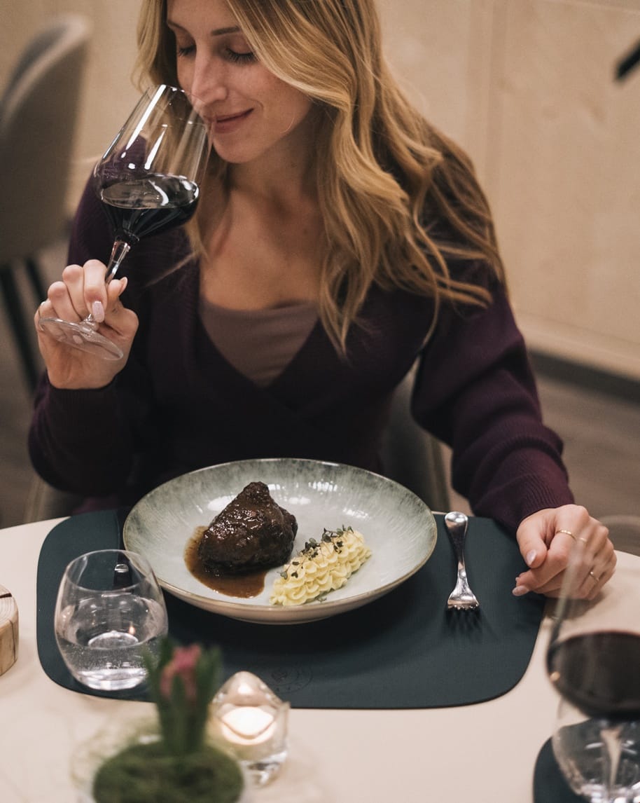 Donna che annusa un calice di vino rosso, seduta a tavola davanti a un piatto con carne e purè