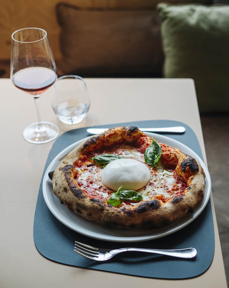 Pizza napoletana con burrata e basilico su piatto, bicchiere di vino e posate su tavolo