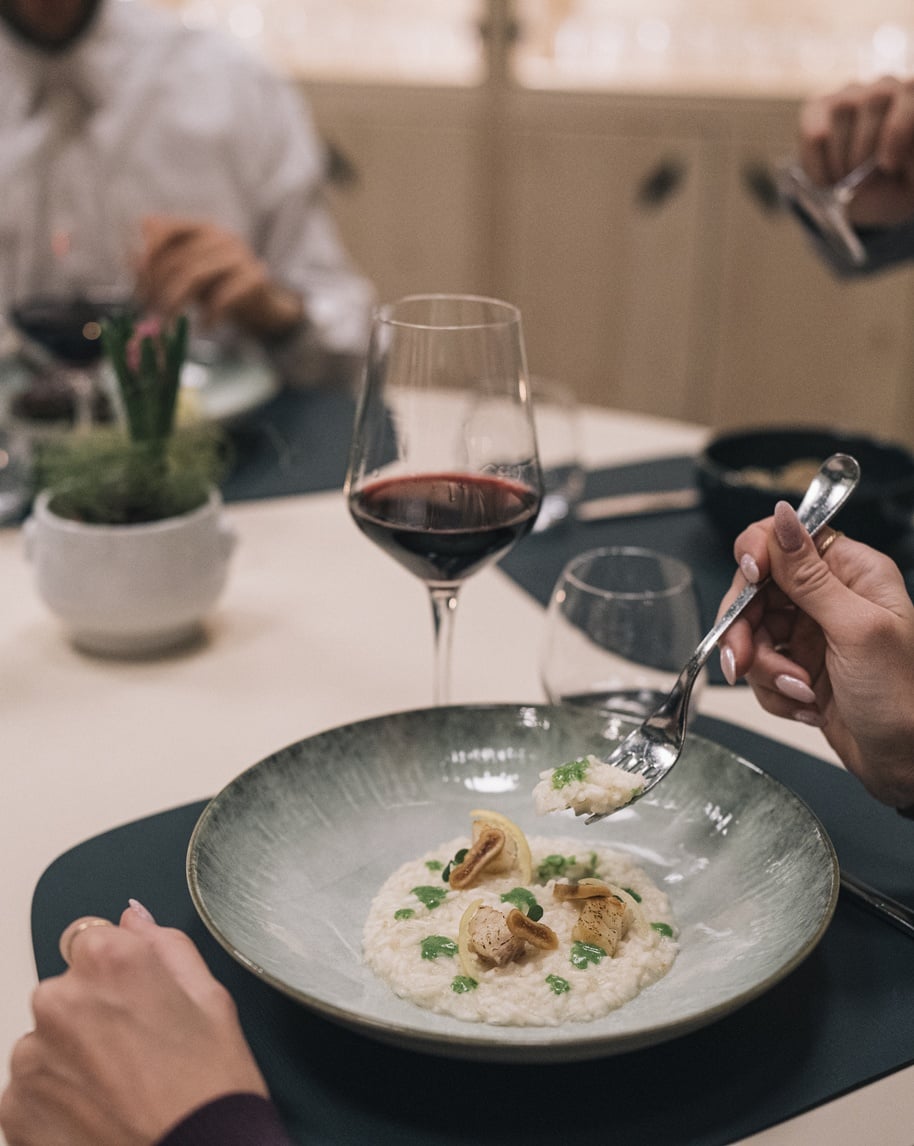 Piatto di risotto con capesante e gocce di salsa verde, mano porta forchettata; calice di vino rosso sul tavolo