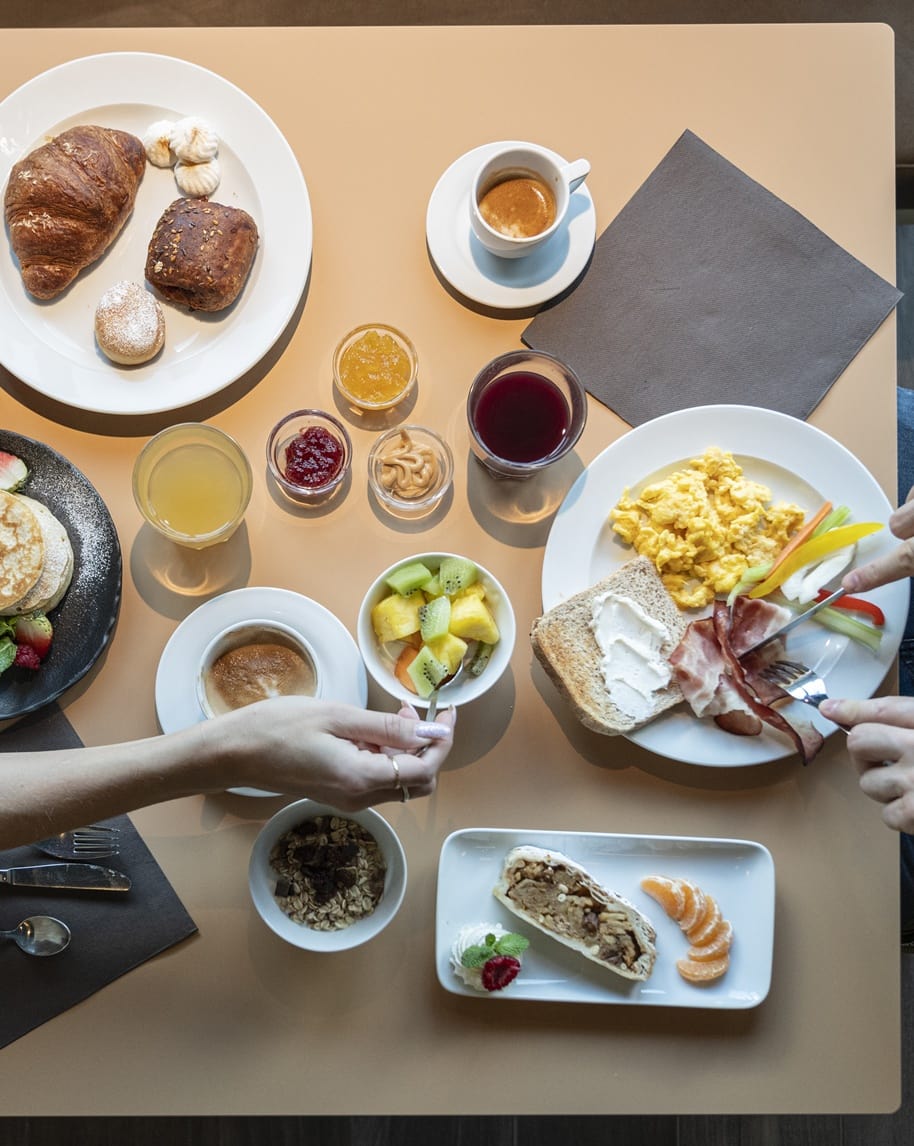 Tavola con colazione: piatti di uova strapazzate, pane, prosciutto, croissant, frutta, caffè