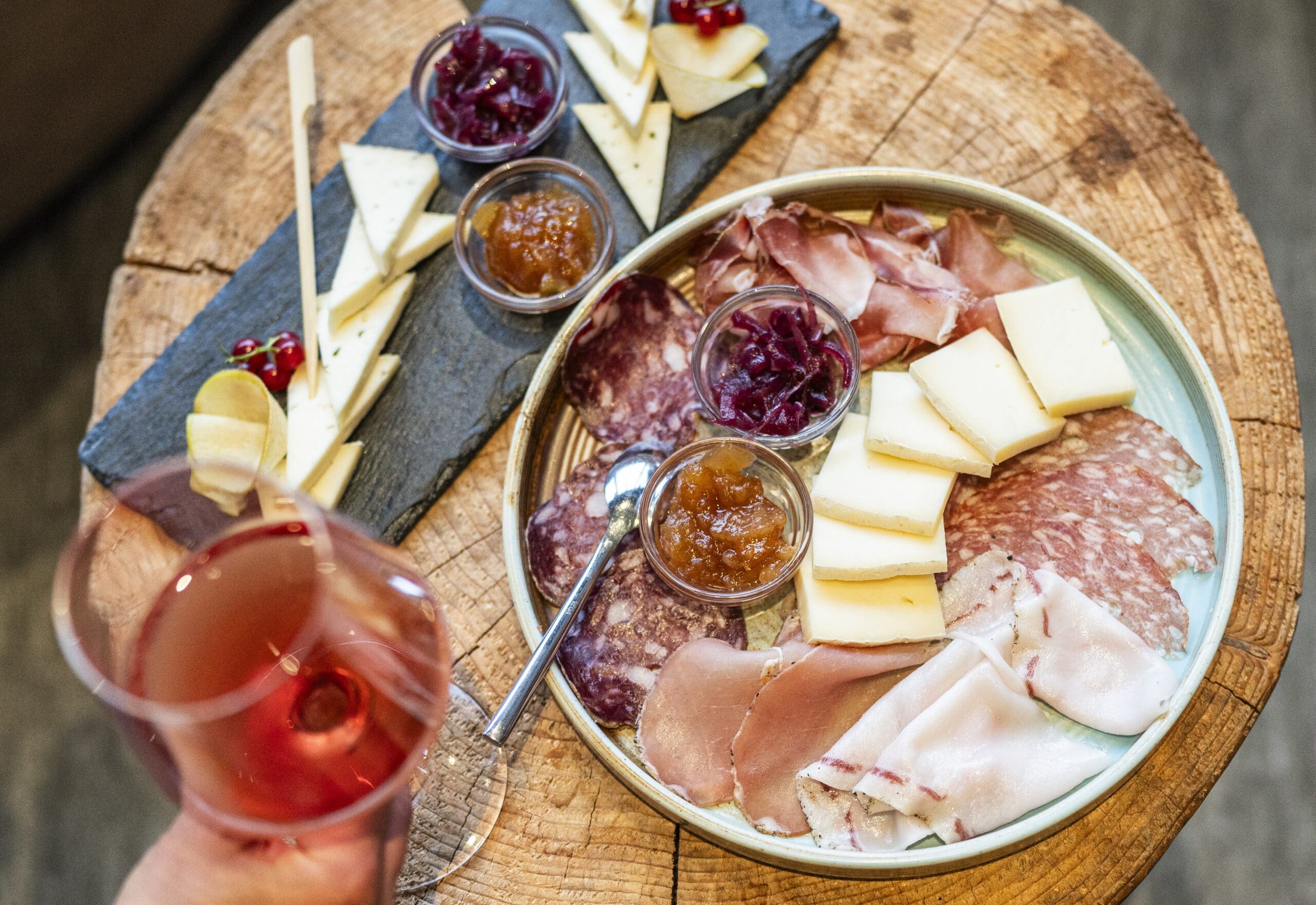 Piatto rotondo con salumi, formaggi e vasetti di conserve su tronco di legno; mano con calice di vino rosato