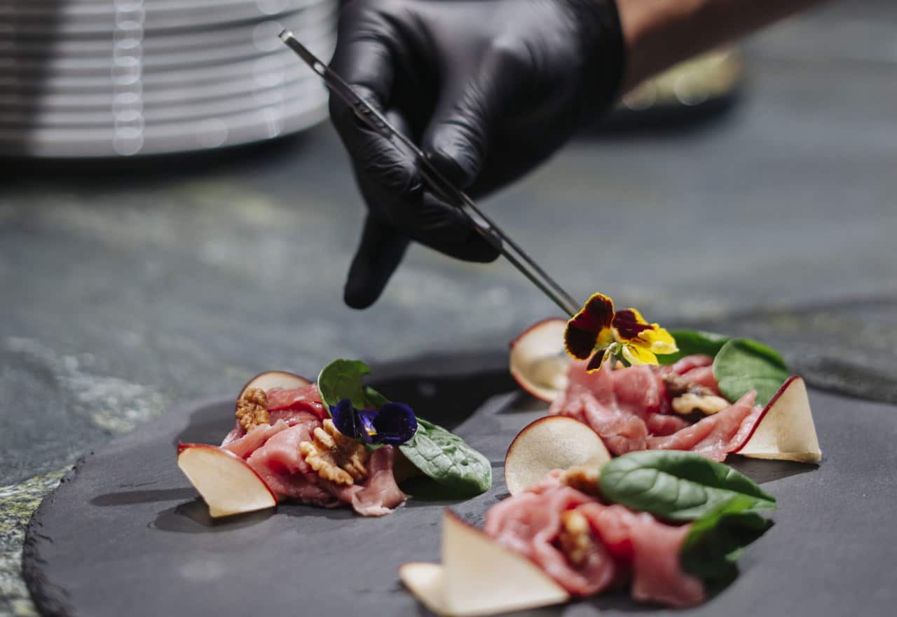 Mano guantata nera sistema fiore commestibile su carpaccio con noci, foglie di spinaci e fette di pera su ardesia