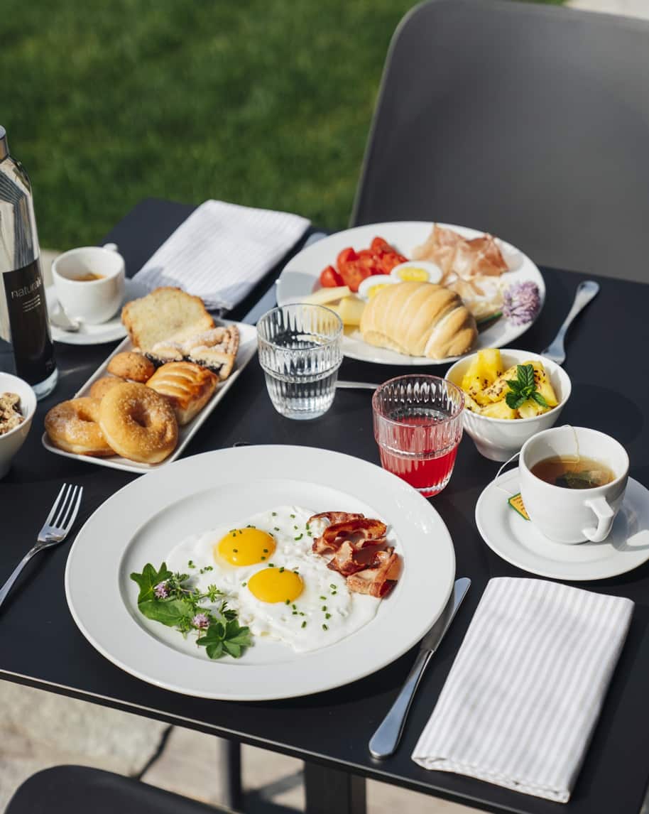 Colazione su tavolo all'aperto: uova con bacon su piatto, brioche, frutta, caffè e tè.