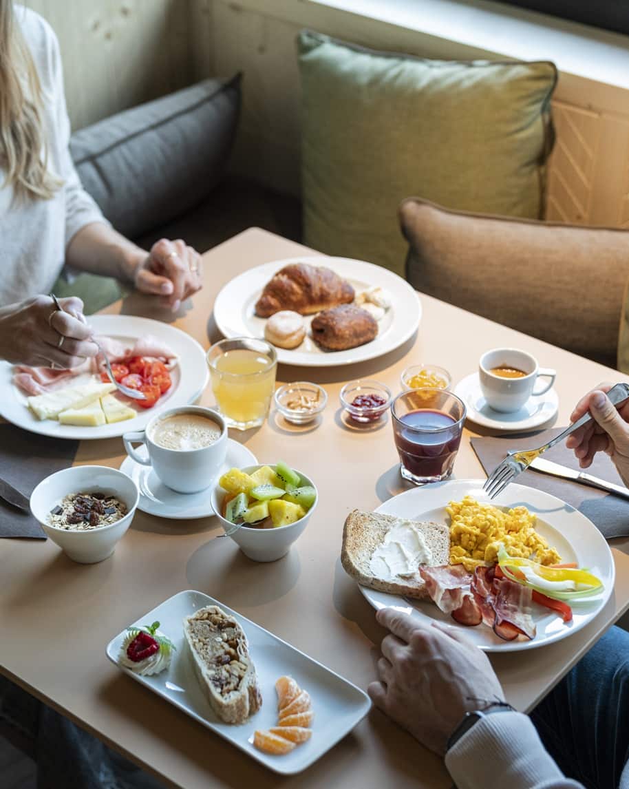 Tavolo con colazione: uova strapazzate, pane, salumi, ciotole di frutta, cornetti, caffè