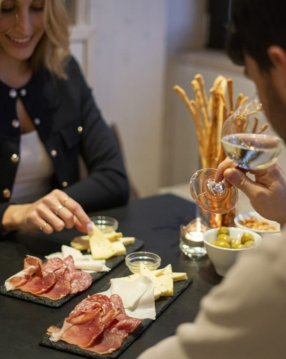Due persone condividono taglieri di salumi e formaggi; una mano tiene un calice di vino bianco.