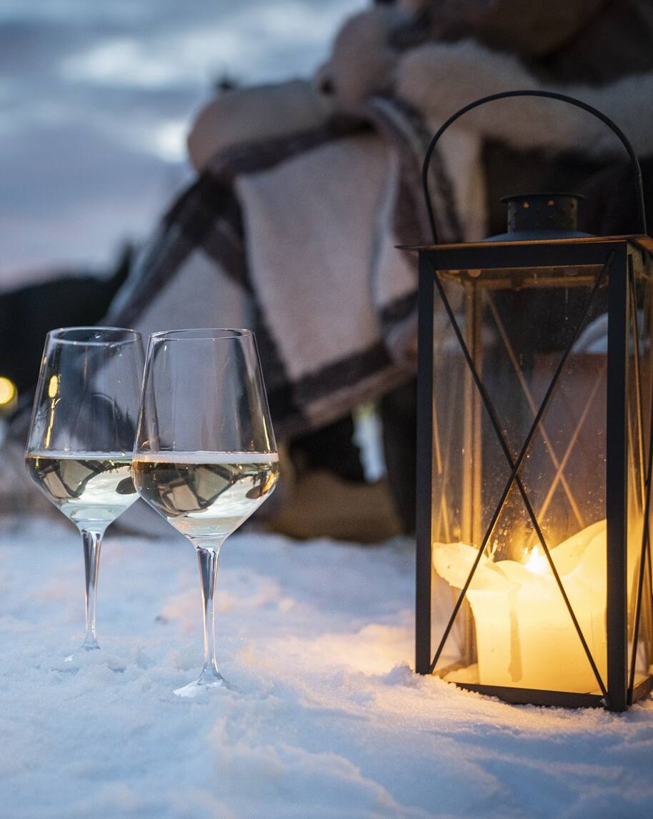 Lanterna con candela accesa accanto a due calici di vino bianco sulla neve, plaid sullo sfondo