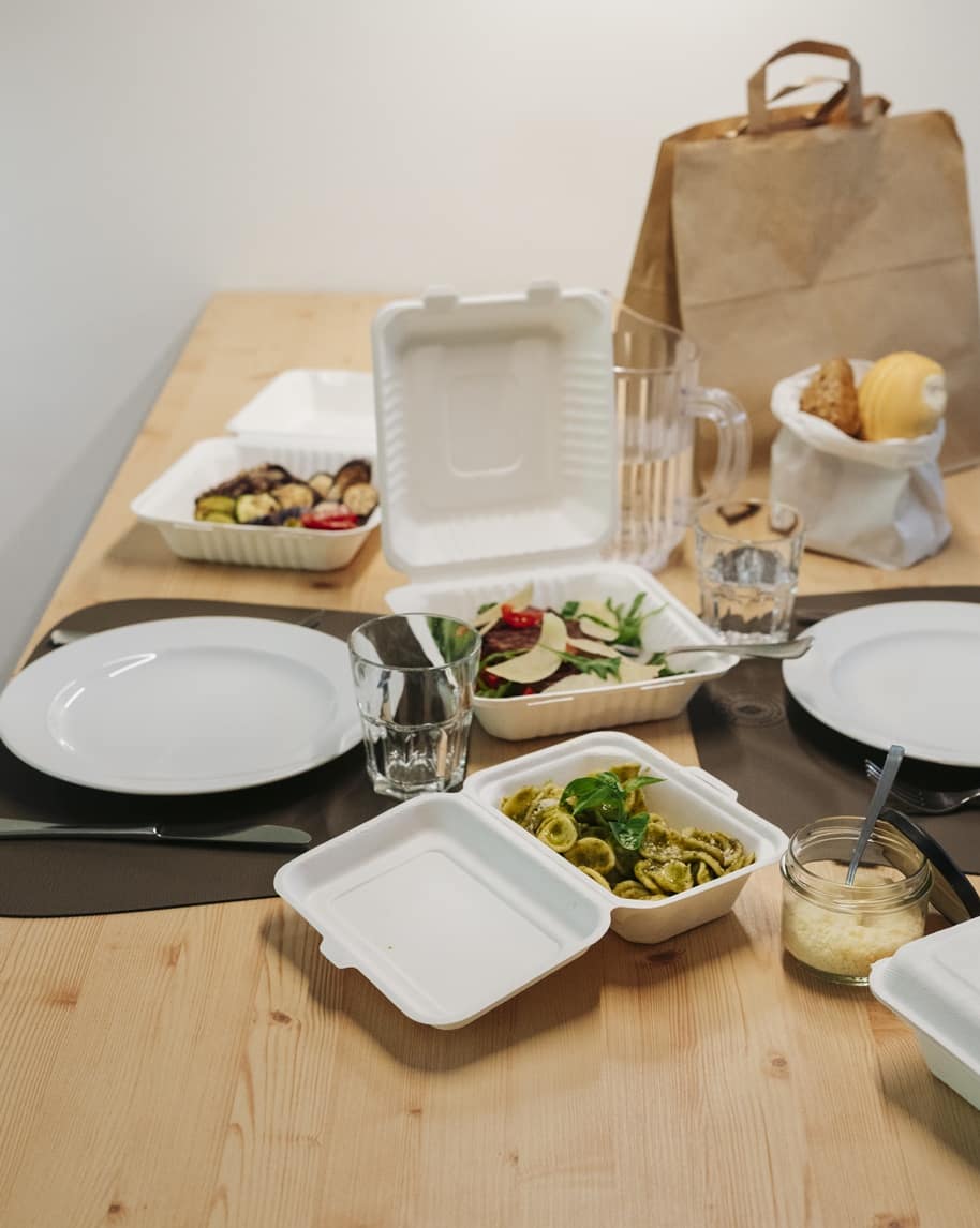Tavolo con contenitori da asporto: pasta al pesto, insalata, piatti, bicchieri e borsa di carta con pane