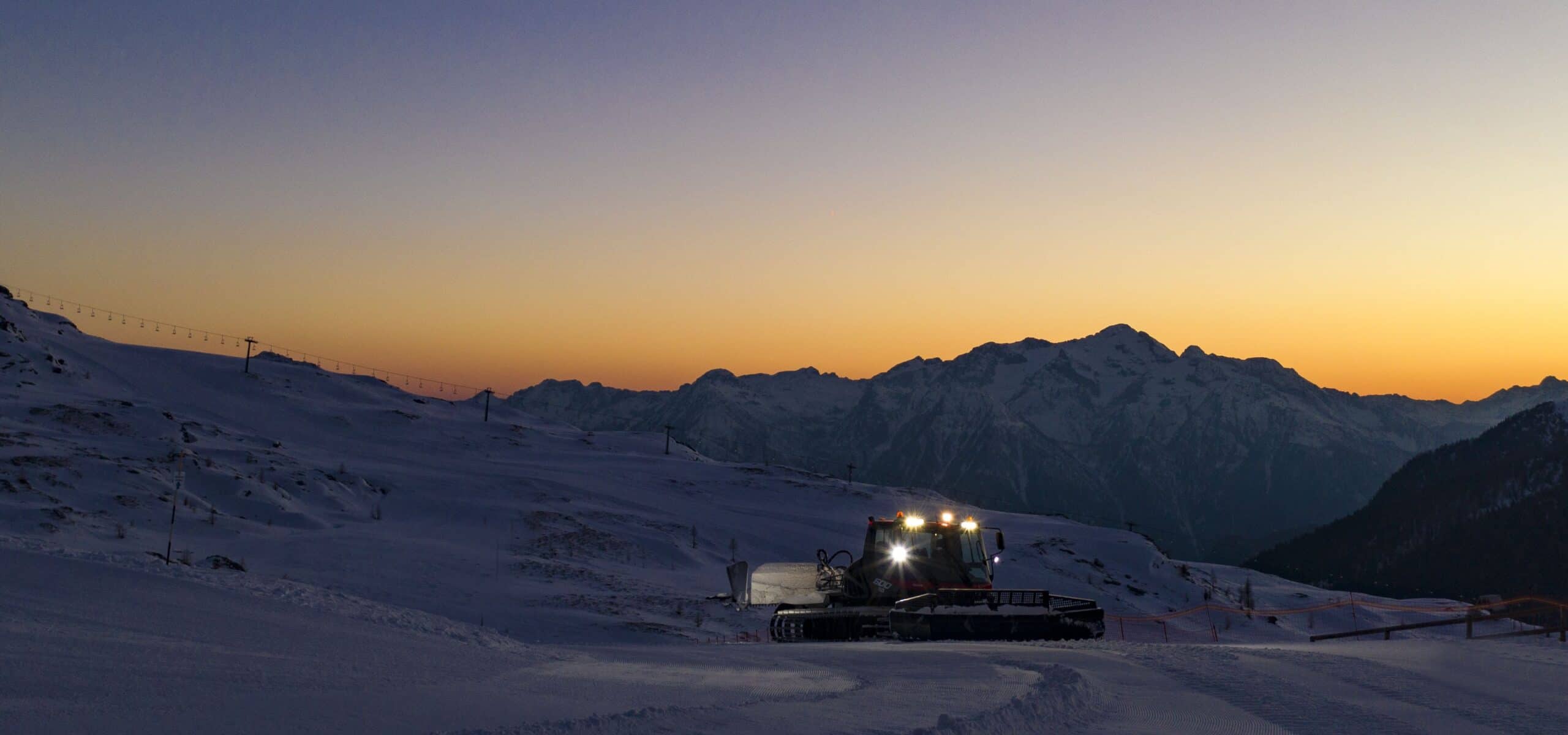 Battipista illuminata su pista innevata al tramonto, pianoro montano con catena di montagne sullo sfondo