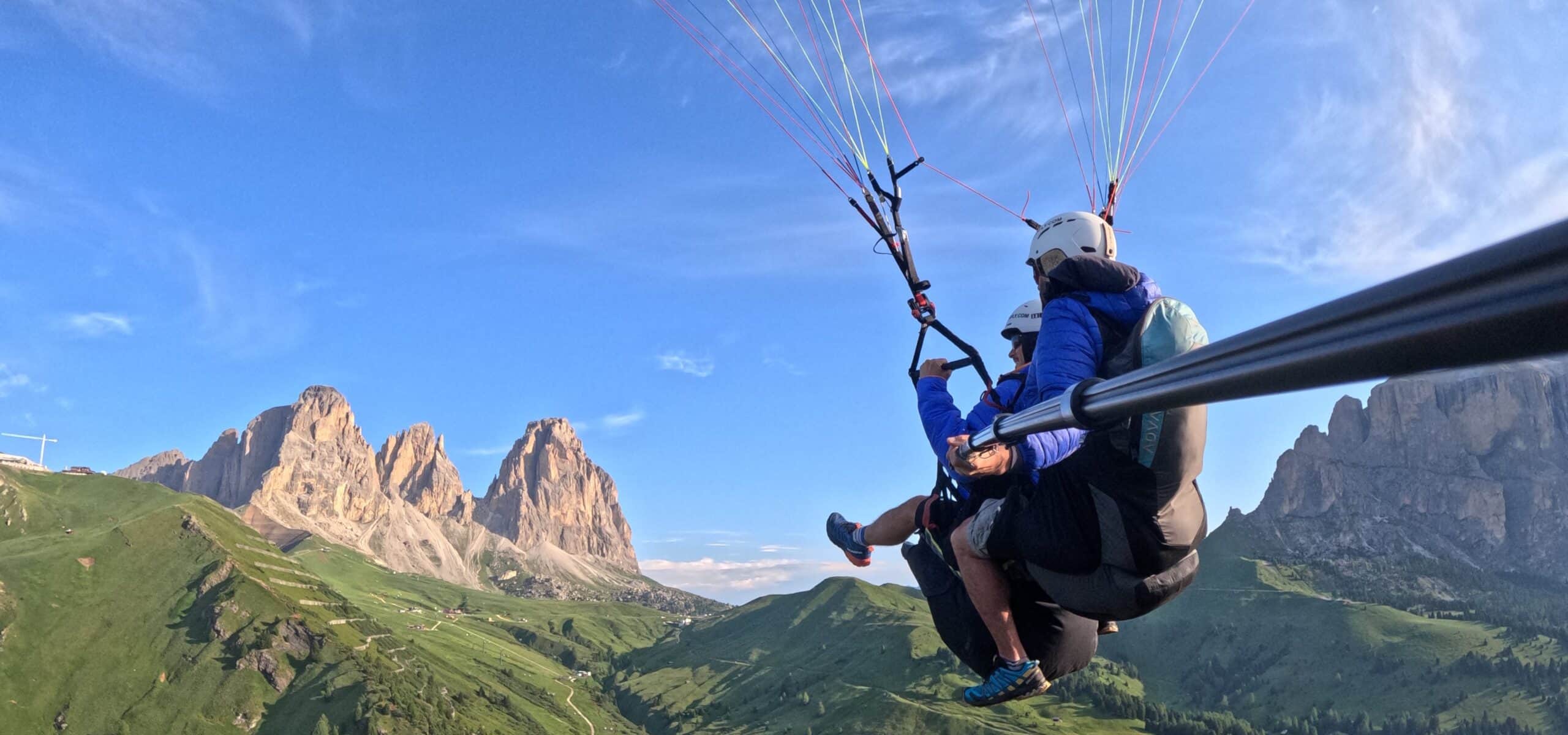 Due persone in parapendio biposto sorvolano una vallata verde con alte pareti rocciose.