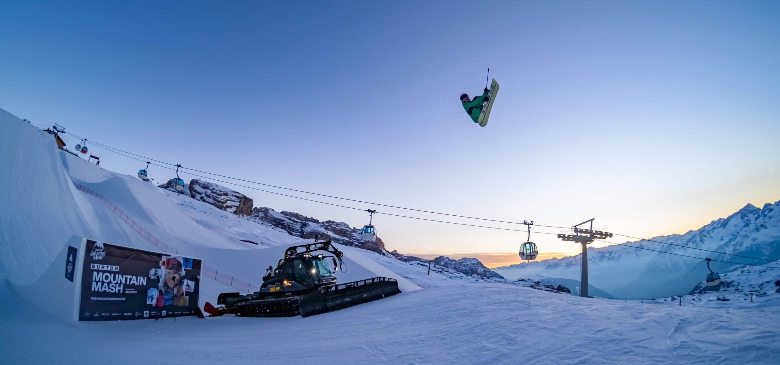 Snowboarder in salto sopra un gatto delle nevi, con cabinovie e montagne innevate sullo sfondo al crepuscolo