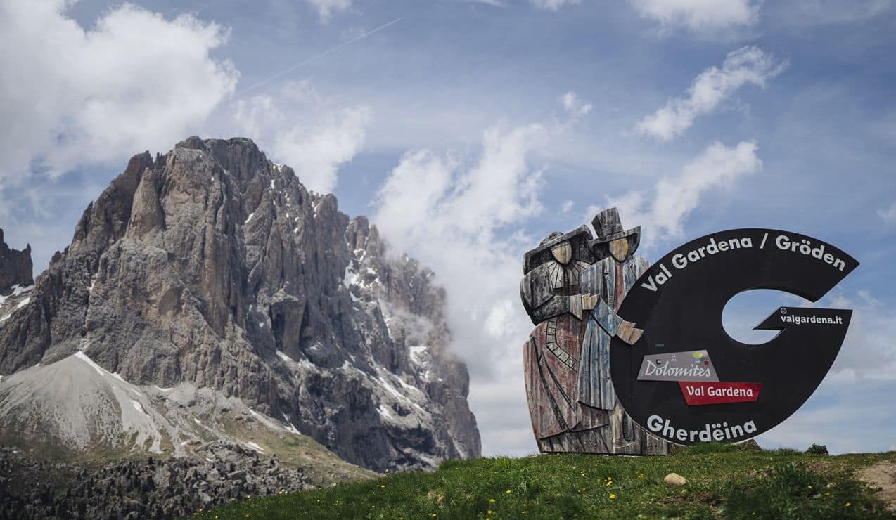 Cartello Val Gardena con sagoma di due figure in legno su prato, parete rocciosa delle Dolomiti sullo sfondo.