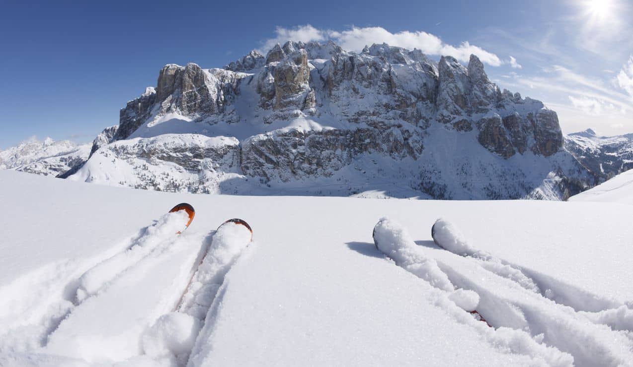 Due paia di sci infossati nella neve, davanti a una massiccia parete rocciosa innevata e cielo sereno