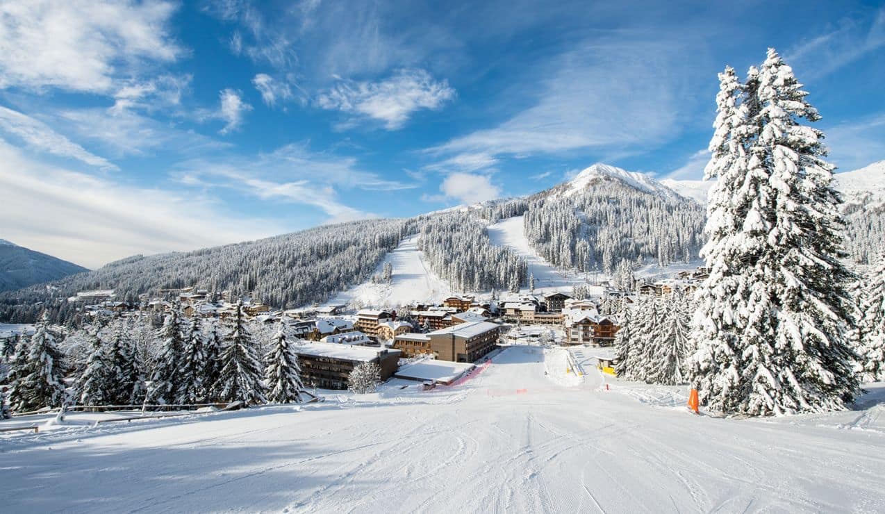 Pista da sci innevata che scende verso un borgo alpino circondato da pini coperti di neve e montagne con tracciati sci
