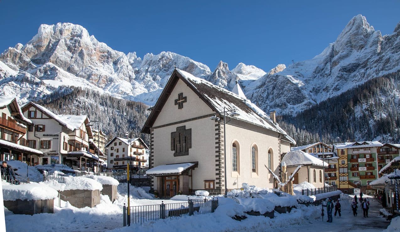 Chiesa al centro coperta di neve, case alpine e persone a piedi davanti, montagne innevate sullo sfondo