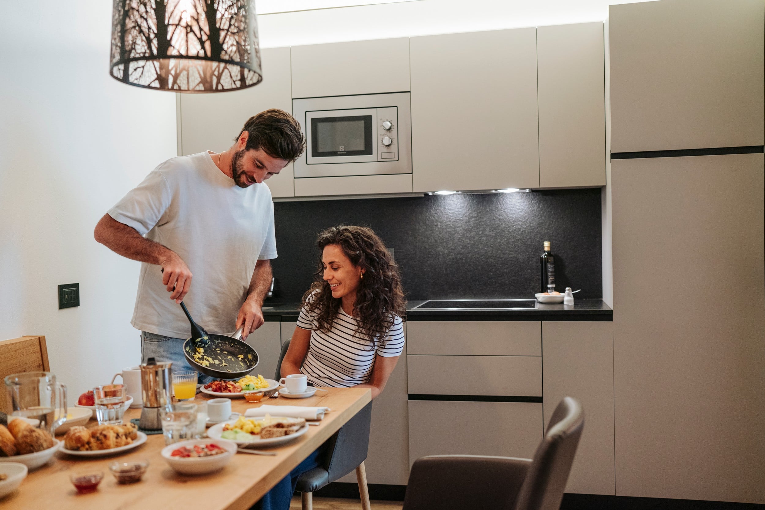Coppia in cucina, uomo versa uova strapazzate da padella nel piatto mentre donna è seduta al tavolo con colazione