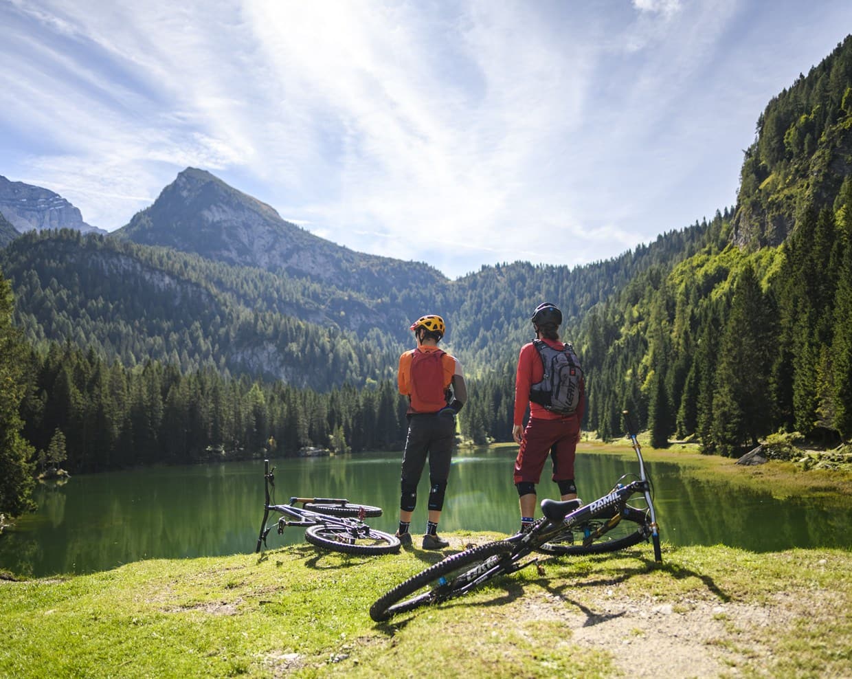 Due ciclisti con caschi e zaini in riva a un lago alpino, biciclette appoggiate sull'erba